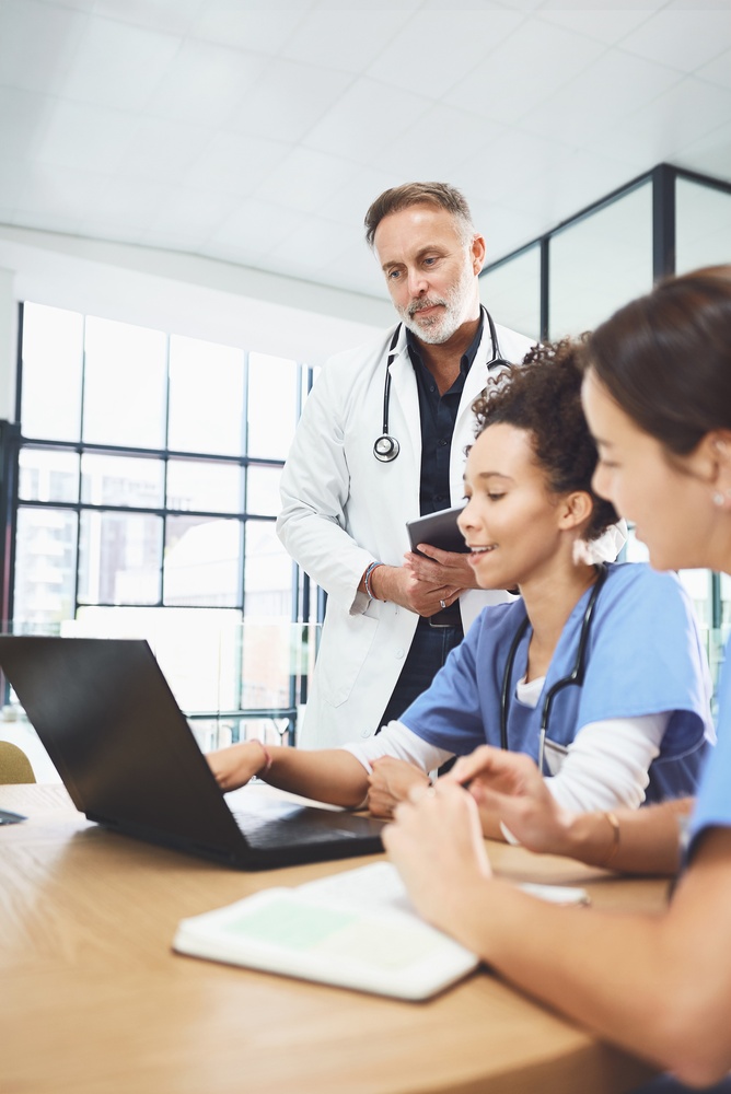 Two men and two women in lab coats with stethoscopes sitting at table and using modern gadgets, afro lady giving papers for colleagues. Multiracial doctors analyzing diagnosis of severe patient.<br />